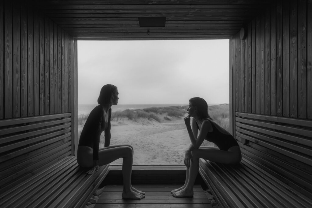 Two women relax in swimsuits with a serene sea view from a wooden sauna.